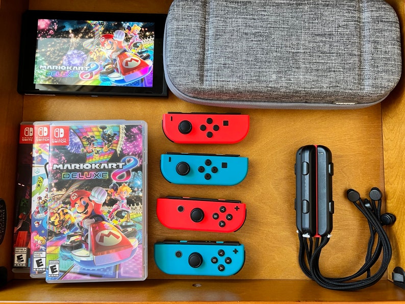 K&ouml;nnte beinhalten: Eine Nintendo Switch-Konsole mit dem Spiel "Mario Kart 8 Deluxe" auf dem Bildschirm und in einer Spielh&uuml;lle. Rote und blaue Joy-Con-Controller, eine graue Tragetasche und ein schwarzer Controller sind ebenfalls zu sehen.