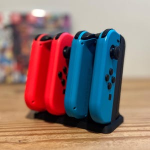 Könnte beinhalten: Eine schwarze Ladestation hält vier farbige Gamecontroller. Zwei rote und zwei blaue Controller sind sichtbar. Die Controller sind für eine Spielkonsole. Die Station steht auf einer Holzoberfläche.