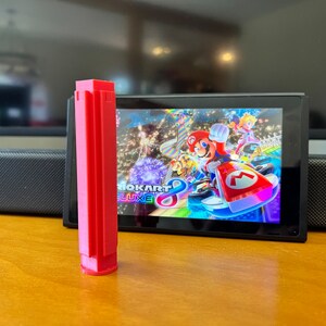 K&ouml;nnte beinhalten: Ein leuchtend rotes, rechteckiges 3D-gedrucktes Objekt steht vor einer Nintendo Switch-Konsole, die das Spiel Mario Kart 8 Deluxe zeigt. Das Spiel zeigt Mario und andere Charaktere beim Rennen. Das Objekt befindet sich auf einer Holzoberfl&auml;che.