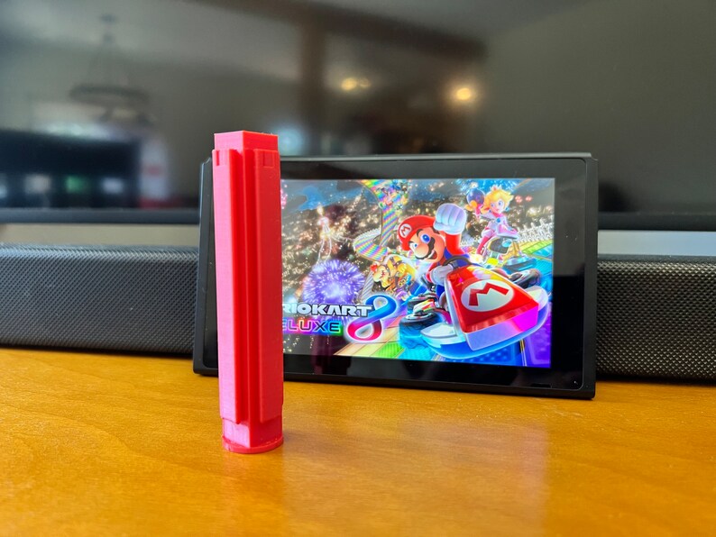 K&ouml;nnte beinhalten: Ein leuchtend rotes, rechteckiges 3D-gedrucktes Objekt steht vor einer Nintendo Switch-Konsole, die das Spiel Mario Kart 8 Deluxe zeigt. Das Spiel zeigt Mario und andere Charaktere beim Rennen. Das Objekt befindet sich auf einer Holzoberfl&auml;che.