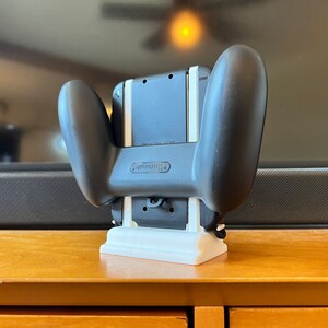 K&ouml;nnte beinhalten: Schwarzer Nintendo Switch Controller-Griff mit wei&szlig;er 3D-gedruckter Basis. Der Controller ist an einer schwarzen Konsole befestigt. Das Nintendo-Logo ist sichtbar. Das Setup befindet sich auf einer Holzoberfl&auml;che.