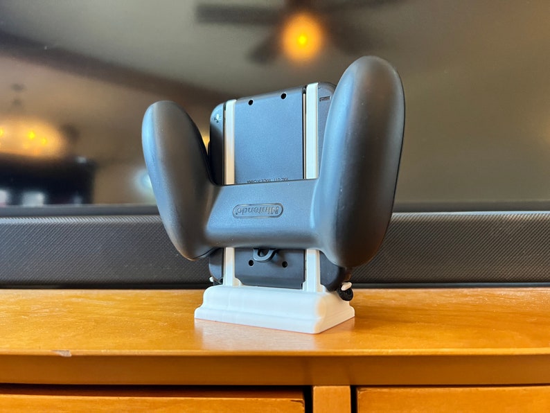 K&ouml;nnte beinhalten: Schwarzer Nintendo Switch Controller-Griff mit wei&szlig;er 3D-gedruckter Basis. Der Controller ist an einer schwarzen Konsole befestigt. Das Nintendo-Logo ist sichtbar. Das Setup befindet sich auf einer Holzoberfl&auml;che.