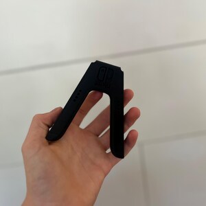 May include: A black plastic stand for a gaming console, held in a hand. The stand has a distinctive A-frame design with the console's logo and four small dots on the side. The stand is designed to hold the console upright.