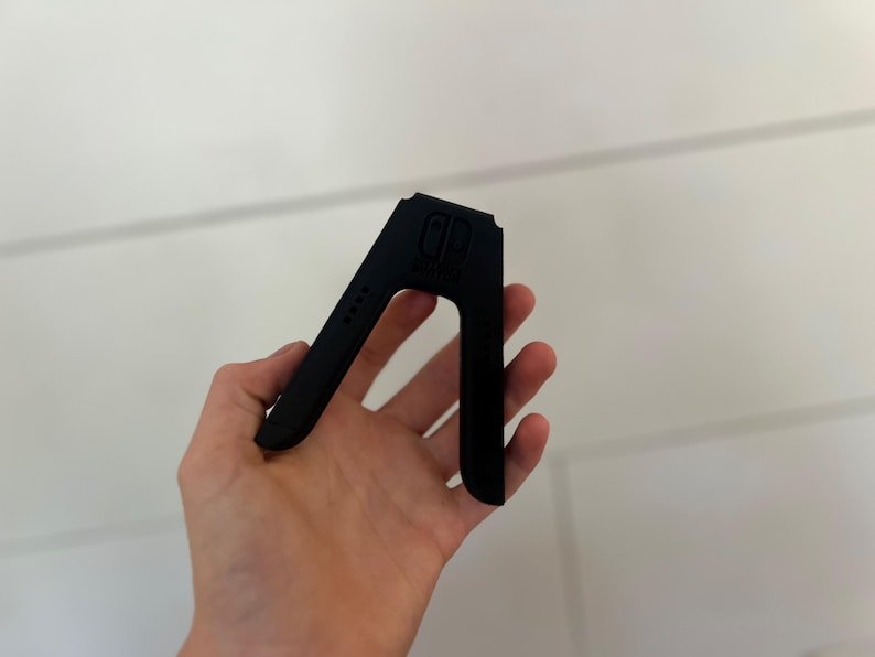 May include: A black plastic stand for a gaming console, held in a hand. The stand has a distinctive A-frame design with the console's logo and four small dots on the side. The stand is designed to hold the console upright.