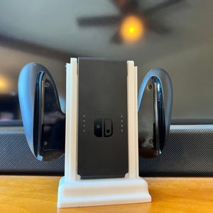 K&ouml;nnte beinhalten: Eine wei&szlig;e und schwarze Nintendo Switch Ladestation. Die Station h&auml;lt die Switch-Konsole und zwei Joy-Con-Controller. Das Switch-Logo ist auf der Konsole sichtbar. Die Station steht auf einer Holzoberfl&auml;che.