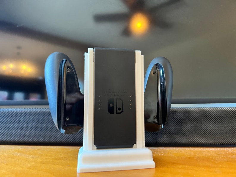 K&ouml;nnte beinhalten: Eine wei&szlig;e und schwarze Nintendo Switch Ladestation. Die Station h&auml;lt die Switch-Konsole und zwei Joy-Con-Controller. Das Switch-Logo ist auf der Konsole sichtbar. Die Station steht auf einer Holzoberfl&auml;che.