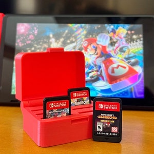 May include: A red Nintendo Switch console with attached Joy-Con controllers, displaying a Mario Kart game. In the foreground, a red case holds three game cartridges, including "Mario Kart 8 Deluxe" and "Minecraft Dungeons Hero Edition".