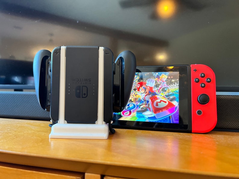 K&ouml;nnte beinhalten: Eine schwarze Nintendo Switch-Konsole mit roten Joy-Con-Controllern, die ein farbenfrohes Rennspiel zeigt. Die Konsole befindet sich in einem wei&szlig;en St&auml;nder, mit einer schwarzen Ladestation dahinter. Der Bildschirm zeigt Mario Kart-Charaktere.