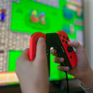 May include: A person holding a red and black video game controller with a green screen in the background. The controller has buttons and a joystick. The screen displays a pixelated video game.