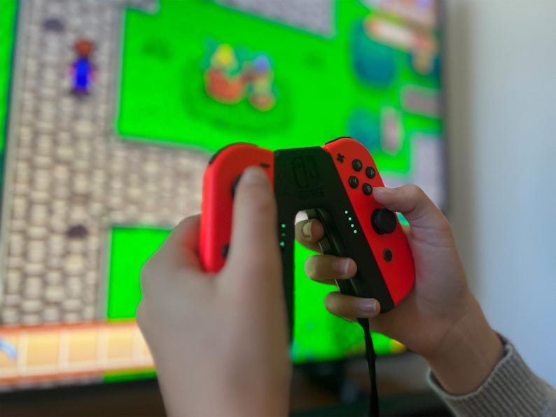 May include: A person holding a red and black video game controller with a green screen in the background. The controller has buttons and a joystick. The screen displays a pixelated video game.