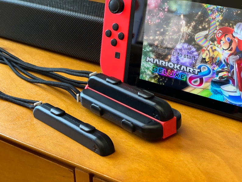 K&ouml;nnte beinhalten: Eine rot-schwarze Nintendo Switch-Konsole mit einem Spielbildschirm, auf dem "Mario Kart 8 Deluxe" angezeigt wird. Zwei schwarze Controller-Zubeh&ouml;rteile mit schwarzen Riemen befinden sich im Vordergrund. Die Konsole und das Zubeh&ouml;r stehen auf einer Holzoberfl&auml;che.