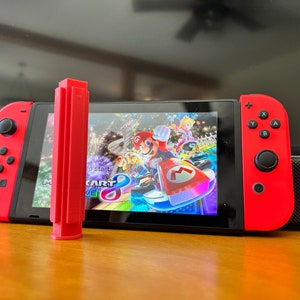 K&ouml;nnte beinhalten: Eine rote Nintendo Switch-Konsole mit roten Joy-Con-Controllern. Der Bildschirm zeigt das Spiel Mario Kart 8 Deluxe. Ein rotes, rechteckiges Zubeh&ouml;r befindet sich vor dem Bildschirm.