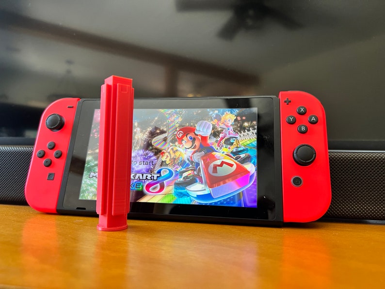 K&ouml;nnte beinhalten: Eine rote Nintendo Switch-Konsole mit roten Joy-Con-Controllern. Der Bildschirm zeigt das Spiel Mario Kart 8 Deluxe. Ein rotes, rechteckiges Zubeh&ouml;r befindet sich vor dem Bildschirm.