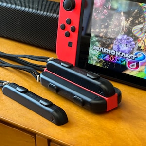 May include: A red and black Nintendo Switch console with a game screen displaying "Mario Kart 8 Deluxe." Two black controller accessories with black straps are in the foreground. The console and accessories are on a wooden surface.