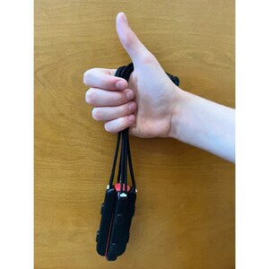 K&ouml;nnte beinhalten: Schwarzes und rotes elektronisches Ger&auml;t mit einem Schl&uuml;sselband, das mit einem Daumen hoch gehalten wird. Das Ger&auml;t hat eine rechteckige Form mit abgerundeten Ecken und einem roten Akzent. Der Hintergrund ist eine Holzoberfl&auml;che.