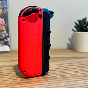 May include: A red and black Nintendo Switch Joy-Con controller. The controller has a black grip and a red body. A game case with the title "Super Smash Bros. Ultimate" is visible in the background.