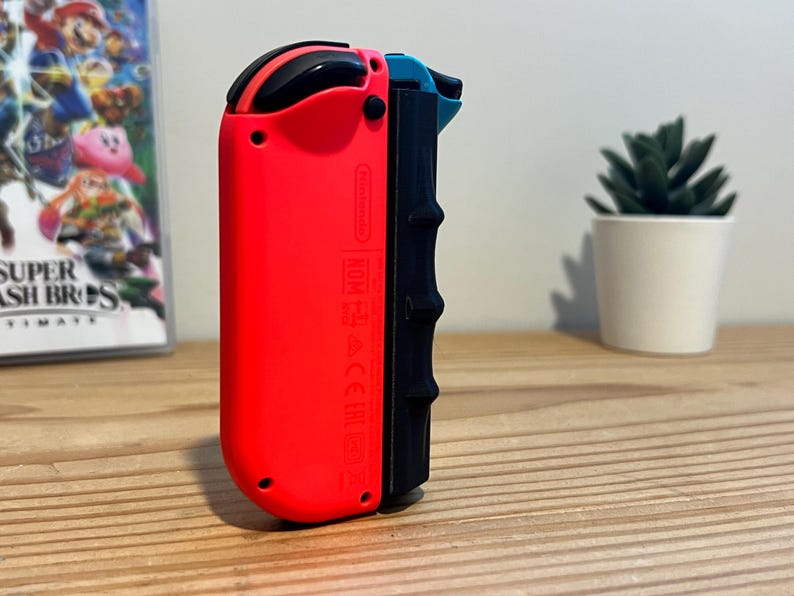 May include: A red and black Nintendo Switch Joy-Con controller. The controller has a black grip and a red body. A game case with the title "Super Smash Bros. Ultimate" is visible in the background.