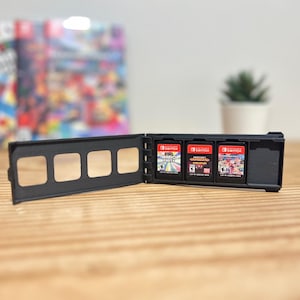 May include: A black Nintendo Switch game cartridge holder with three game cartridges visible. The cartridges are red and black with game titles. A blurred game box and a small potted plant are in the background.