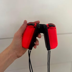 May include: A pair of red and black game controllers, held in a hand. The controllers have a sleek design with black buttons and a black connecting piece. Black wrist straps hang from the bottom of each controller.
