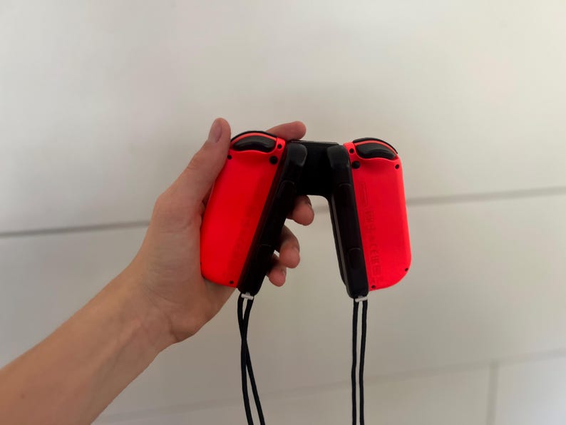 May include: A pair of red and black game controllers, held in a hand. The controllers have a sleek design with black buttons and a black connecting piece. Black wrist straps hang from the bottom of each controller.