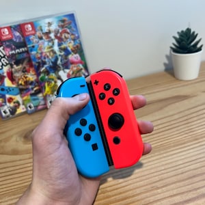 May include: A pair of red and blue Nintendo Switch Joy-Con controllers are held in a hand. The controllers are in front of two game cases with colorful graphics. A small potted plant is in the background.