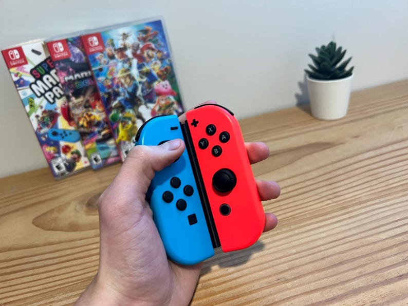 May include: A pair of red and blue Nintendo Switch Joy-Con controllers are held. The controllers are in front of two game cases with colourful graphics. A small potted plant is in the background.