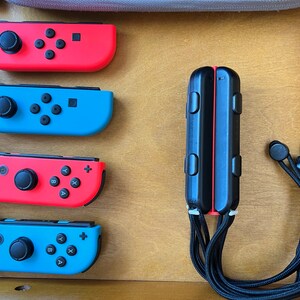 K&ouml;nnte beinhalten: Eine Sammlung von Nintendo Switch-Zubeh&ouml;r, darunter rote und blaue Joy-Con-Controller, ein schwarzer Controller-Griff und schwarze Ohrh&ouml;rer. Die Controller sind auf einer Holzoberfl&auml;che angeordnet.