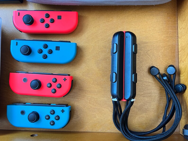 K&ouml;nnte beinhalten: Eine Sammlung von Nintendo Switch-Zubeh&ouml;r, darunter rote und blaue Joy-Con-Controller, ein schwarzer Controller-Griff und schwarze Ohrh&ouml;rer. Die Controller sind auf einer Holzoberfl&auml;che angeordnet.
