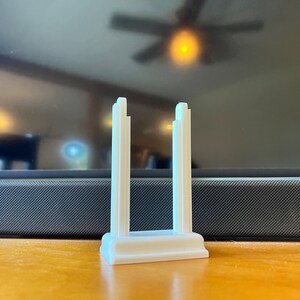 K&ouml;nnte beinhalten: Ein wei&szlig;es, 3D-gedrucktes Objekt, m&ouml;glicherweise ein St&auml;nder oder Halter, mit rechteckiger Basis und zwei vertikalen, architektonisch gestalteten St&uuml;tzen. Die St&uuml;tzen haben ein abgestuftes Design. Das Objekt befindet sich auf einer Holzoberfl&auml;che.