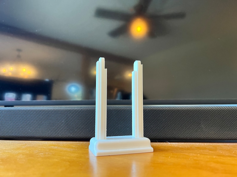 K&ouml;nnte beinhalten: Ein wei&szlig;es, 3D-gedrucktes Objekt, m&ouml;glicherweise ein St&auml;nder oder Halter, mit rechteckiger Basis und zwei vertikalen, architektonisch gestalteten St&uuml;tzen. Die St&uuml;tzen haben ein abgestuftes Design. Das Objekt befindet sich auf einer Holzoberfl&auml;che.