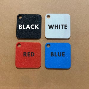 K&ouml;nnte beinhalten: Vier quadratische, strukturierte Anh&auml;nger in Schwarz, Wei&szlig;, Rot und Blau. Jeder Anh&auml;nger hat oben ein kleines Loch und ein wei&szlig; gedrucktes Wort: BLACK, WHITE, RED und BLUE. Die Anh&auml;nger sind auf einer braunen Oberfl&auml;che angeordnet.