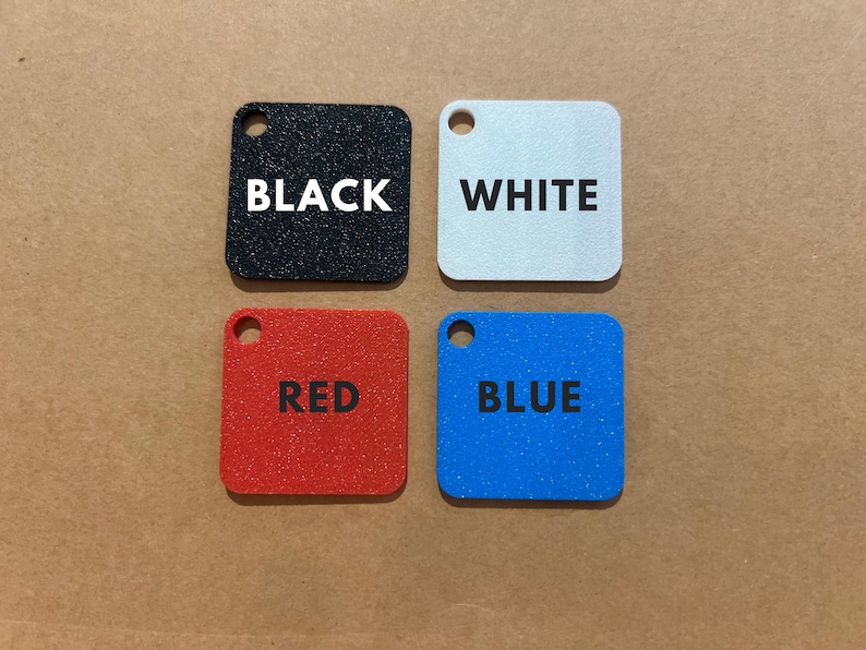 K&ouml;nnte beinhalten: Vier quadratische, strukturierte Anh&auml;nger in Schwarz, Wei&szlig;, Rot und Blau. Jeder Anh&auml;nger hat oben ein kleines Loch und ein wei&szlig; gedrucktes Wort: BLACK, WHITE, RED und BLUE. Die Anh&auml;nger sind auf einer braunen Oberfl&auml;che angeordnet.