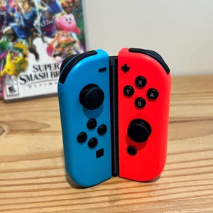 May include: A pair of detached blue and red Nintendo Switch Joy-Con controllers. The blue controller has a black joystick and black buttons. The red controller has a black joystick and black buttons. The controllers are on a wooden surface.