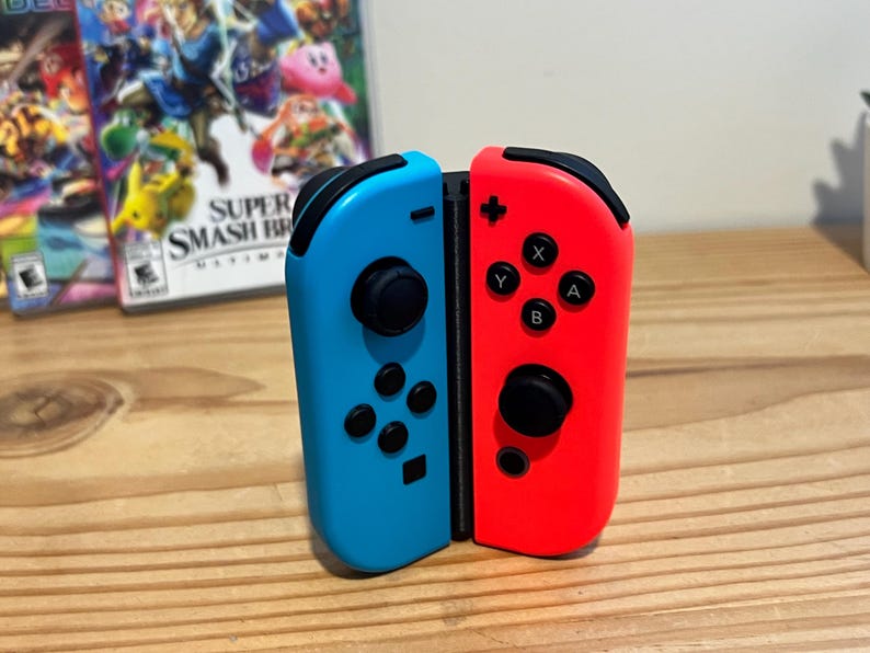 May include: A pair of detached blue and red Nintendo Switch Joy-Con controllers. The blue controller has a black joystick and black buttons. The red controller has a black joystick and black buttons. The controllers are on a wooden surface.
