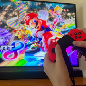 May include: A person holding red and black video game controllers in front of a television screen displaying the Mario Kart 8 Deluxe game. The game features Mario and Princess Peach in a colorful racing scene.