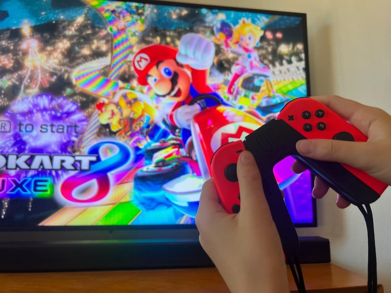 May include: A person holding red and black video game controllers in front of a television screen displaying the Mario Kart 8 Deluxe game. The game features Mario and Princess Peach in a colorful racing scene.