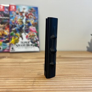 May include: A black, vertical, 3D-printed object with three raised, geometric shapes along its side. The object is on a wooden surface, with three video game cases in the background. A small potted plant is also visible.