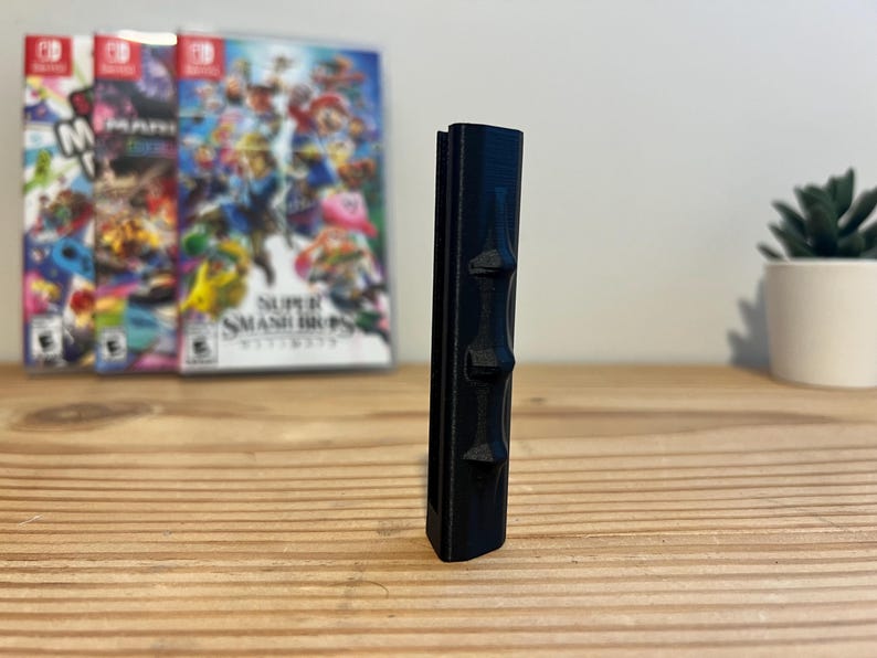 May include: A black, vertical, 3D-printed object with three raised, geometric shapes along its side. The object is on a wooden surface, with three video game cases in the background. A small potted plant is also visible.