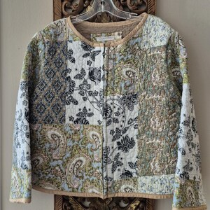 May include: A quilted jacket featuring a patchwork design with various patterns in shades of blue, green, black, and white. The garment has a round neckline, long sleeves, and a front zip closure, finished with solid tan trim along the edges.