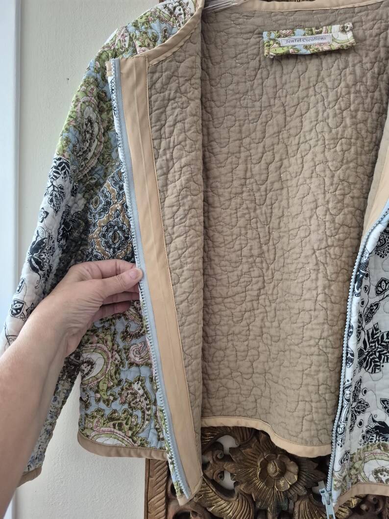 Pu&ograve; includere: Una giacca trapuntata con cerniera caratterizzata da un motivo floreale e paisley multicolore nei toni del blu, verde, nero e bianco. L'interno &egrave; foderato con tessuto beige tinta unita e include un'etichetta con la scritta Tenfel Creations.