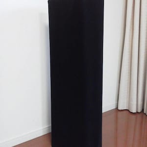 以下が含まれることがあります： 白い壁を背景に、角が丸い長方形の黒い物体が立っています。物体は柔らかく暗い素材でできているようです。背景には木の床と白いカーテンがあります。