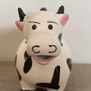 Peut inclure: Un pot à crème en céramique blanche en forme de vache avec des taches noires et des oreilles roses. La vache a un nez noir et une bouche rose.