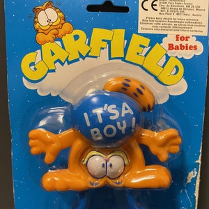 Peut inclure: Un hochet pour bébé Garfield bleu et orange avec le texte "It's a Boy!" sur la partie bleue. Le hochet est emballé sur une carte bleue et blanche avec le logo Garfield et le texte "for Babies".