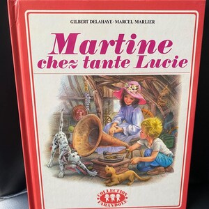 Peut inclure: Un livre cartonné rouge avec le titre "Martine chez tante Lucie" en grandes lettres blanches. La couverture présente une illustration d'une fille, d'un garçon et d'un chien dans une pièce avec un gramophone. Le livre fait partie de la collection "Collection Farandole" et est publié par Casterman.