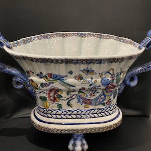 Puede incluir: Un gran cuenco de cerámica blanca con diseños florales azules y dorados y motivos de pájaros. El cuenco tiene dos asas con forma de cabeza de pájaro y se apoya en tres patas.