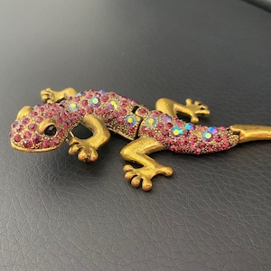 Könnte beinhalten: Eine goldfarbene, mit rosa und schillernden Strasssteinen besetzte Gecko-Figur. Der Gecko ist in einer Gehposition mit eingerolltem Schwanz dargestellt.