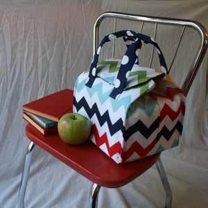 Peut inclure: Un sac &agrave; d&eacute;jeuner color&eacute; avec un motif en chevron rouge, blanc, bleu, vert et jaune. Le sac a une poign&eacute;e noire avec des pois blancs. Le sac est pos&eacute; sur une chaise rouge avec une pile de livres et une pomme verte.