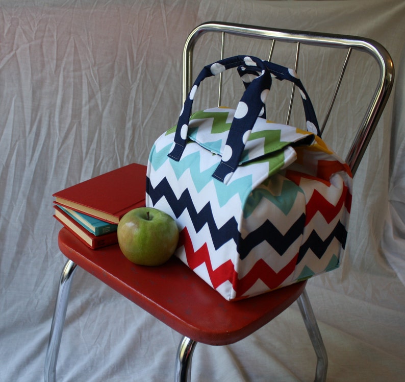 Peut inclure: Un sac &agrave; d&eacute;jeuner color&eacute; avec un motif en chevron rouge, blanc, bleu, vert et jaune. Le sac a une poign&eacute;e noire avec des pois blancs. Le sac est pos&eacute; sur une chaise rouge avec une pile de livres et une pomme verte.