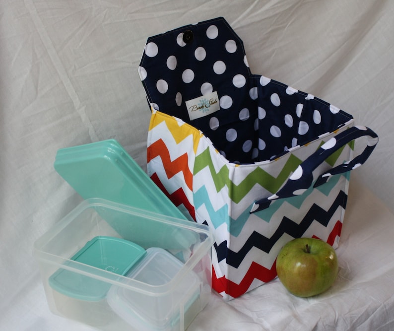 Peut inclure: Un sac &agrave; d&eacute;jeuner color&eacute; avec un motif en chevron et une doublure en pois bleu marine. Le sac a une poign&eacute;e et est ouvert pour r&eacute;v&eacute;ler un r&eacute;cipient &agrave; d&eacute;jeuner en plastique transparent avec un couvercle vert.