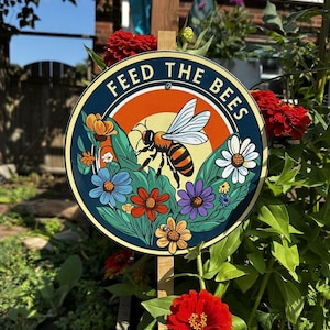 May include: Circular sign displaying "FEED THE BEES" in white text. The sign features a bee illustration, set against a sunset backdrop, surrounded by colorful flowers. It's mounted on a wooden stake, amidst red flowers and green foliage.